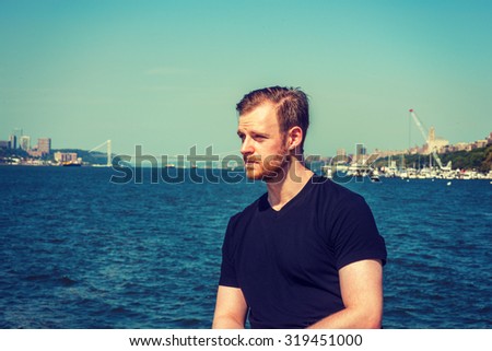 American man traveling in New York. Wearing black v neck T shirt, a young guy with beard, mustache standing by Hudson River, looking forward. Bridge, harbor on background. Instagram effect.