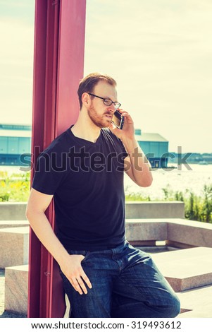 Technology in our daily life. Wearing black v neck T shirt, jeans, a young man with beard, mustache leaning against red pole by Hudson River in New York, relaxing, talking on phone. Instagram effect.