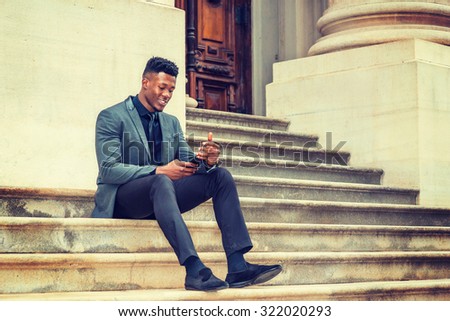 Dressing in fashionable jacket, necktie, pants, cloth shoes, a young black businessman sitting on stairs outside vintage style office building, smiling, checking messages on his cell phone. Texting.