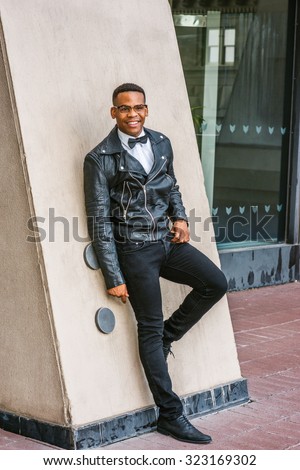 Man Urban Autumn/Spring Casual Fashion. Wearing black leather jacket, jeans, leather shoes, glasses, white undershirt, bow tie, an African American guy standing by wall on street in New York, smiling.