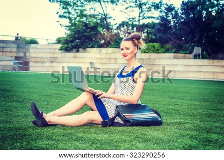 American businesswoman working in New York. A young sexy college student wearing blue, white polkadot dress, carrying leather bag, sitting on green lawn on campus, smiling, working on laptop computer.