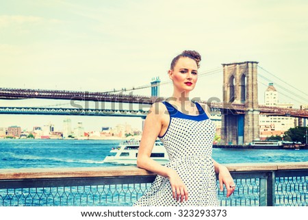 American businesswoman traveling in New York. Wearing blue, white polkadot dress, a young sexy lady standing at harbor by East River, looking at you. Manhattan, Brooklyn bridges, boat on background.