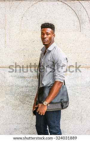 Man on the way going to work. Wearing gray shirt, jeans, wristwatch, carrying shoulder leather bag, an African American guy standing by wall on street, listening music with earphone and cell phone.