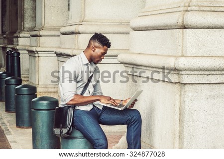 African American College Student studying in New York. Wearing gray shirt, jeans, carrying shoulder leather bag, a black man sitting on metal pillar on street, reading, working on laptop computer.
