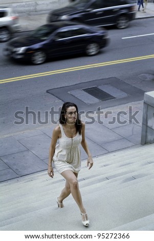 Business Woman Walking up to Work. Dressing in a light yellow sleeveless dress, sandals, a young pretty professional lady is forcefully walking up on stairs to the office. A busy street on background