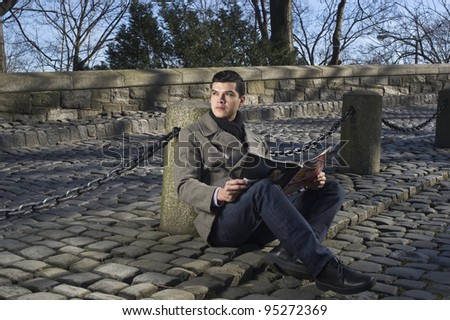 Man Winter Fashion. Dressing in overcoat, black woolen scarf, jeans, leather shoes, a young guy sitting on ground outside against a metal chain fence, reading a magazine, relaxing, waiting for you.