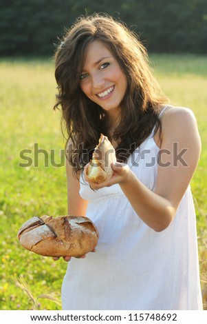 Happy and smiling girl with white country dress is offering a piece of bread toward the camera . Outdoor natural light