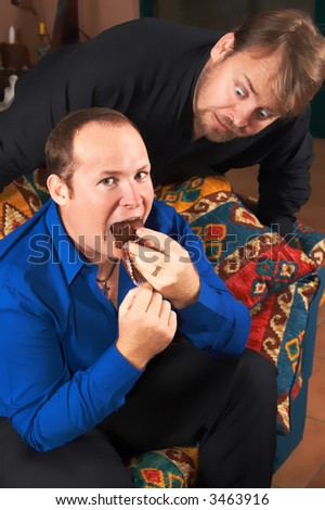 One hungry man staring at the other one eating a huge chocolate muffin.
