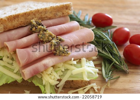 Tasty open sandwich with green lettuce, grated cheese, smoked ham and wholegrain mustard on wholewheat bread with rosemary and tomatoes on chopping board