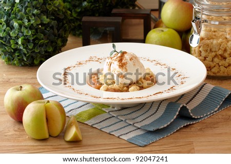apple crumble topped with a scoop of vanilla ice cream decorated with cinnamon powder in a white plate with fresh apples, a jar of crumble and a small green tree background