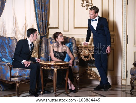 Woman in evening dress and young man in suit sitting in the armchairs and conversing with another man