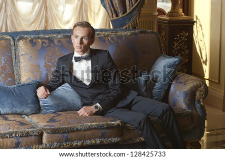 Young blond man in suit and bow tie sitting on blue sofa in old luxury interior