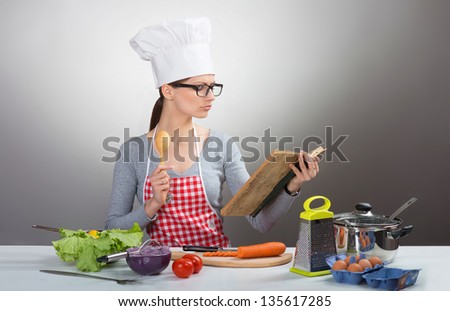 Pretty serious woman cooking with old cookbook on grey background