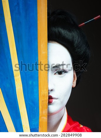 Closeup portrait of a woman in geisha makeup covering half of her face with a big fan