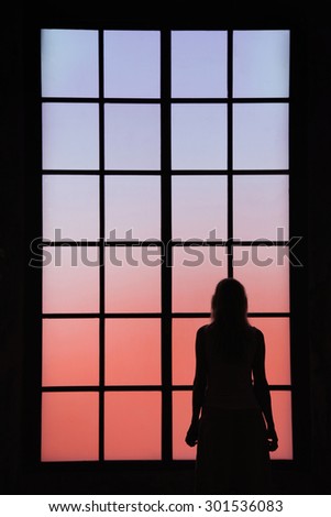 Lonely women looking through big closed glass window and thinking about things. Conceptual lonely or sad women silhouette background.