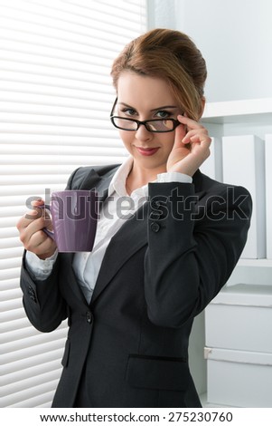 Playful young businesswoman taking off her glasses and looking at camera. Woman standing near the window with a cup of tea during coffee break
