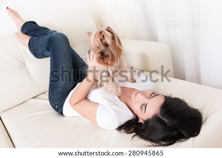Beautiful young brunette girl playing with her yorkshire terrier at home laying down at sofa and holding her pet