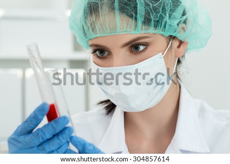 Medical doctor in protective gloves and surgical mask and hat comparing two flasks with dark red liquid in laboratory. Scientific research, healthcare and medical concept.