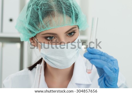 Medical doctor in protective gloves and surgical mask and hat looking at small flask with liquid in laboratory. Scientific research, healthcare and medical concept.