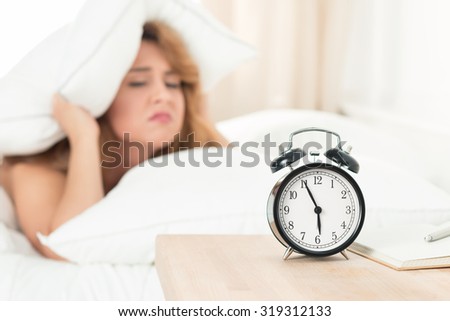 Young beautiful woman hates waking up early in the morning. Sleepy girl looking at alarm clock and trying to hide under the pillow