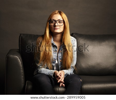 Attractive young woman in glasses sitting on black sofa on black background