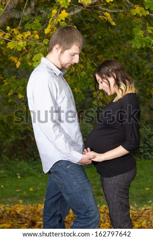 A couple expecting their first child together stand facing each other outside on an Autumn day. They are both looking down gazing at her swollen belly while holding hands.