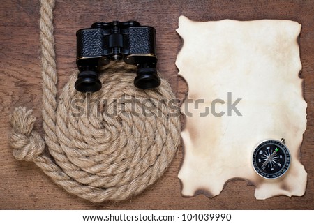 Compass, binoculars, burnt grunge paper and rope on wooden background
