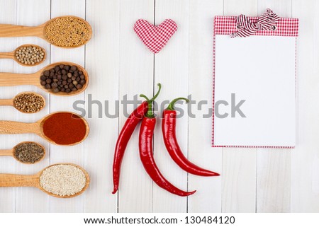 Recipe notebook, heart, chili, spices in spoons on white wood background