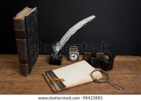 Antique book, magnifying glass, old notepad, quill ink pen and inkwell, vintage binoculars, clock on wood table