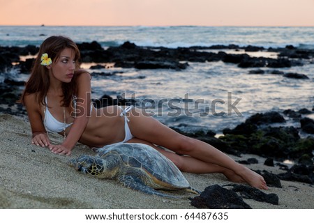 A woman in a  white bikini poses on the beach next to a resting sea turtle