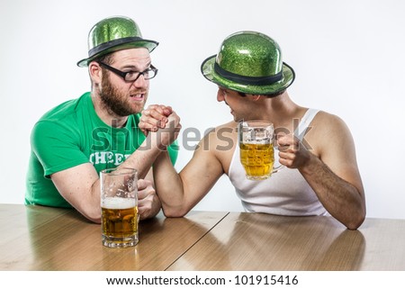 Irish Men arm wrestling in green hats on St. Patty\'s Day with mugs of frothy beer at local tavern, in white tank top and shirt