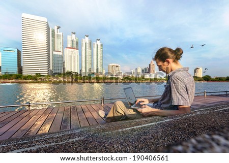 Yang man is working outdoors near river and business office buildings, using a laptop computer