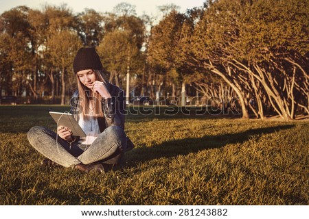 Young student hipster girl with tablet sitting in park, reading and listening to music