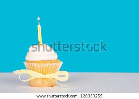 Single vanilla cupcake with lit yellow candle and sprinkles on a blue background and white table