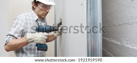 man drywall worker or plasterer using cordless electric screwdriver to fix the plasterboard sheets to the metal profiles to build the new wall . Wearing white hardhat, work gloves and safety glasses