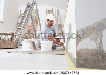 man plasterer construction worker at work, takes plaster from bucket and puts it on trowel to plastering the wall, wears helmet inside the building site of a house Stock fotó © 