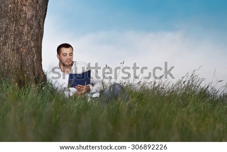 Man lie down on grass and read book. Guy leaning against a tree relaxing - resting. Education in the nature