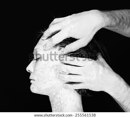 Black and white portrait of human hands working with woman with clay on face on black background