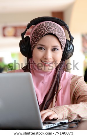 Young pretty Asian muslim woman in head scarf listens to audio with headphone while working on laptop in cafe