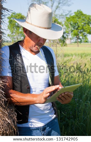 A cowboy leaning on the hay stack in a countryside setting using touchscreen on a tablet computer