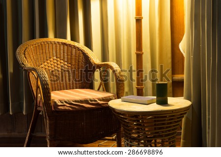 Rattan chair and stool under the light from the lamp with bamboo cup and the book on the stool all surround by beautiful curtain.