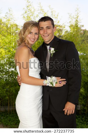 Prom couple outside posing for a photo.  The girl is blond and wearing a beautiful white dress and her date is wearing a black tuxedo with a black shirt.  They are smiling for the camera