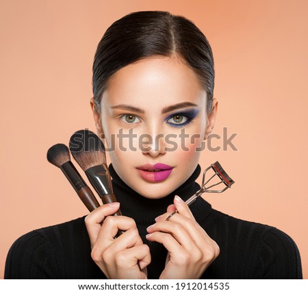 Portrait of a girl with  tools for making makeup near face.  One half face of a beautiful white woman with  bright makeup and the other is natural. Woman holds makeup brush and eyelash curler. Natural