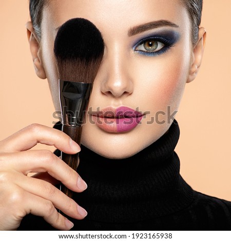 Portrait of a girl with cosmetic brush at face. Woman covering one eye on the face using makeup brush. One half face of a beautiful white woman with  bright makeup and the other is natural.  