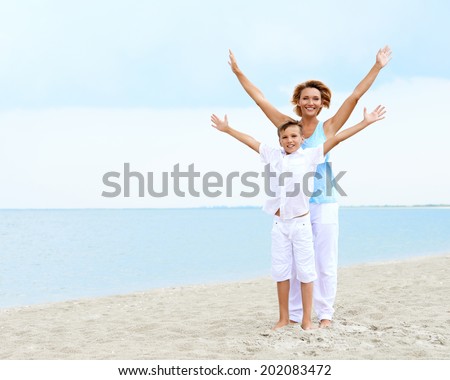Happy smiling mother and son standing on the beach with raised hands.