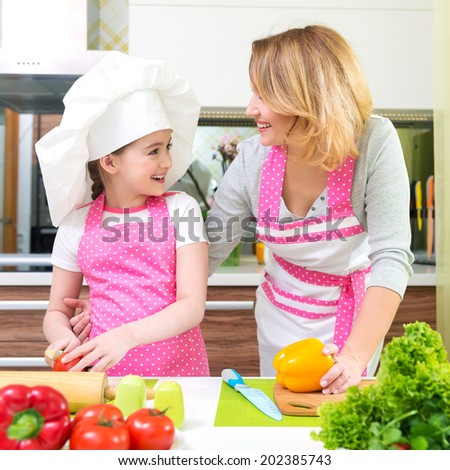 Smiling young mother with daughter in pink apron cooking vegetables at the kitchen.