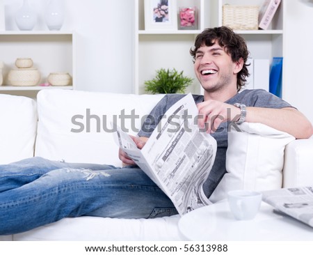 Happy laughing handsome man in casuals lying on the sofa with newspaper - indoors