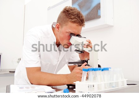 A young male lab technician looks at slides through a microscope whilst comparing the results to earlier tests.