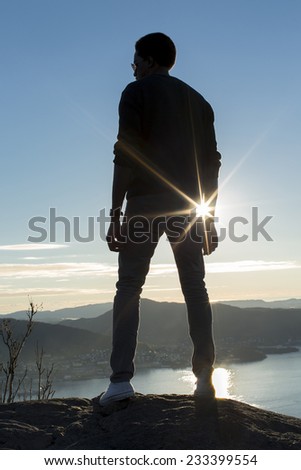 Silhouette male in casual clothes standing atop a mountain with sun flare as the horizon and sea are seen below him, concept of success