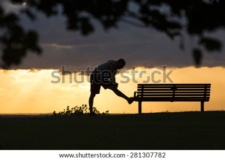 Silhouette male at sunset stretching against a bench